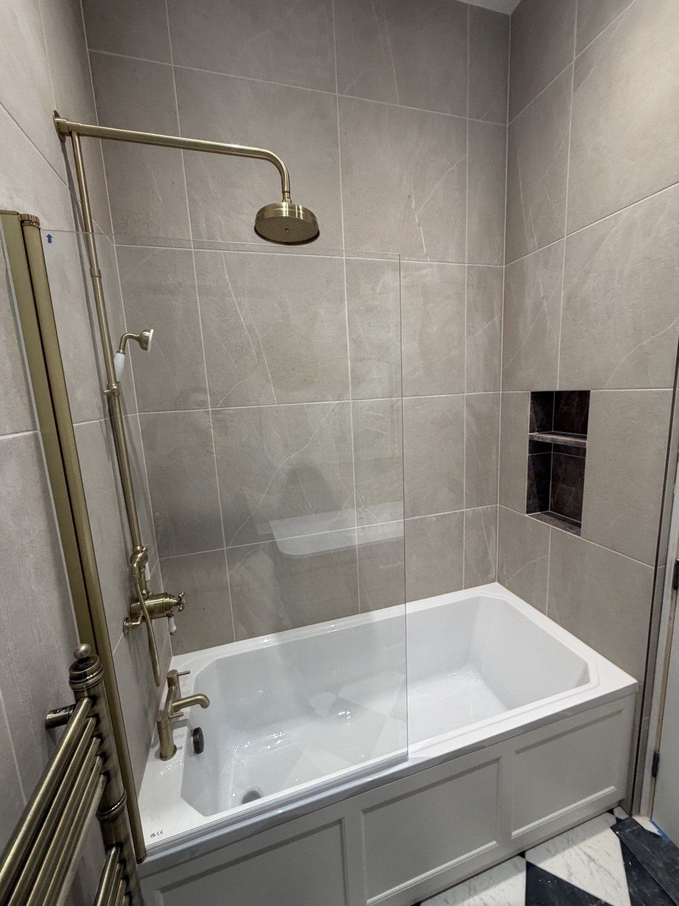 A shower over a bath with an antique-brass exposed shower, large-format stone-effect tiles and a recessed niche